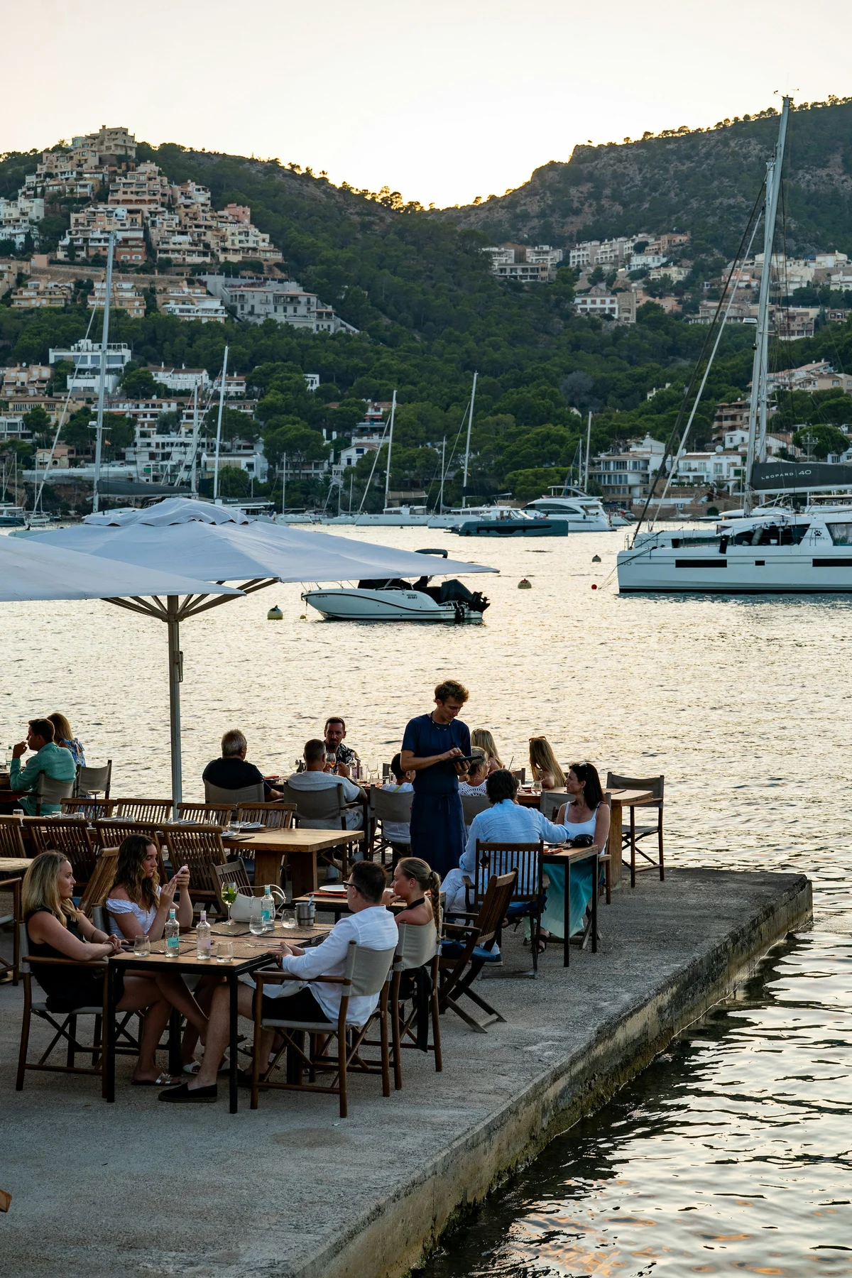 Personas compartiendo mesa junto al agua en Port d’Andratx, Mallorca, con barcos y colinas al fondo.